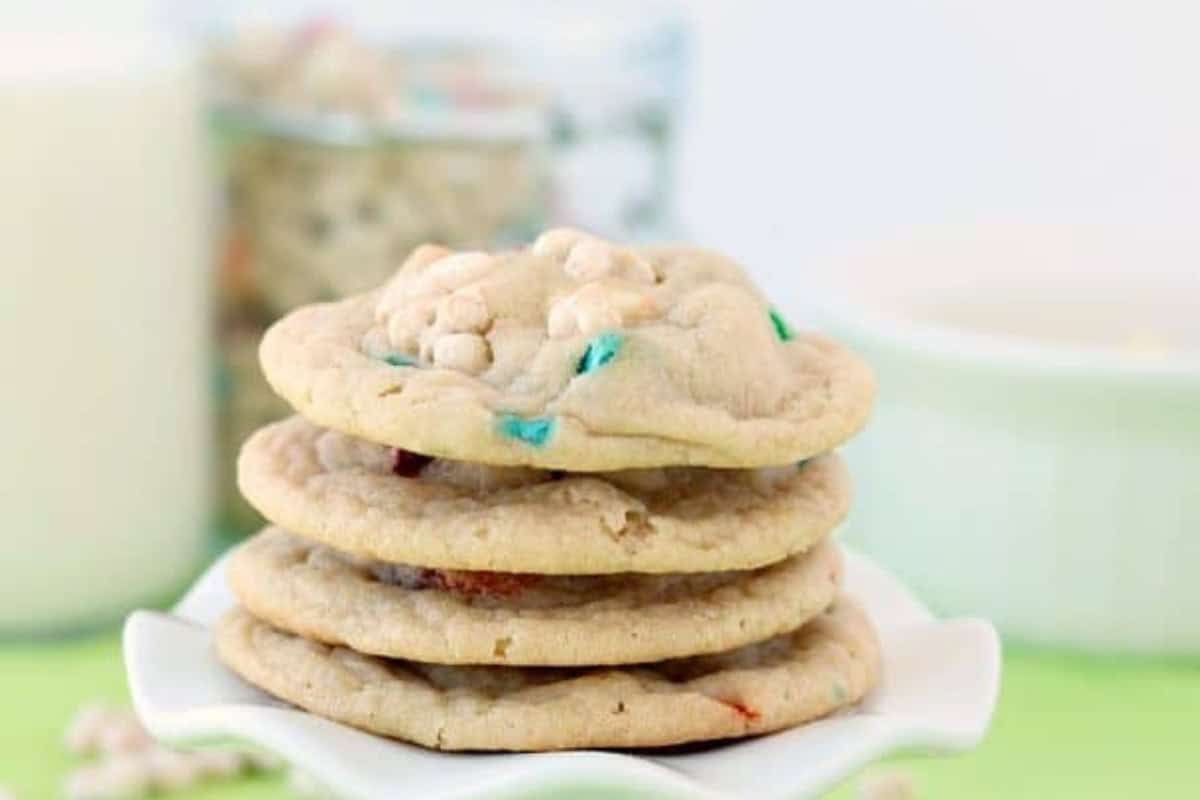 A stack of four cookies with colorful candy pieces sits on a white dish, part of an indulgent desserts spread, with a glass of milk and a jar in the blurred background.