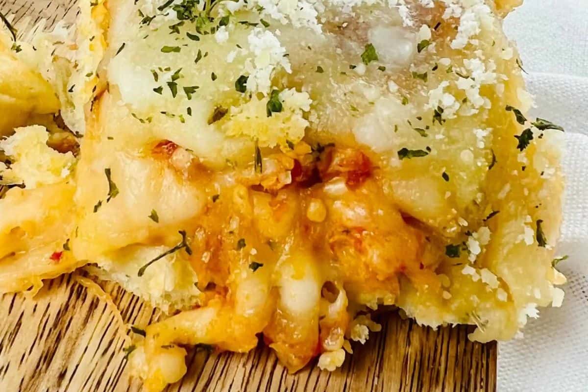 A close-up of a slider with a cheesy, saucy meatball filling, topped with melted white cheese and sprinkled with green herbs and grated cheese. The slider rests on a wooden surface.