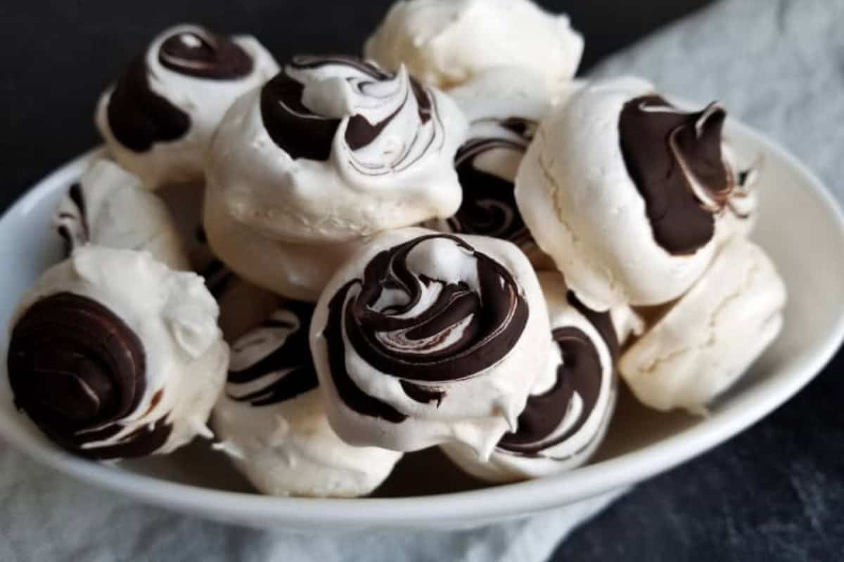 A white bowl filled with small meringue cookies, each topped with a swirl of dark chocolate—perfect for anyone seeking indulgent desserts or adding to their chocolate collection.