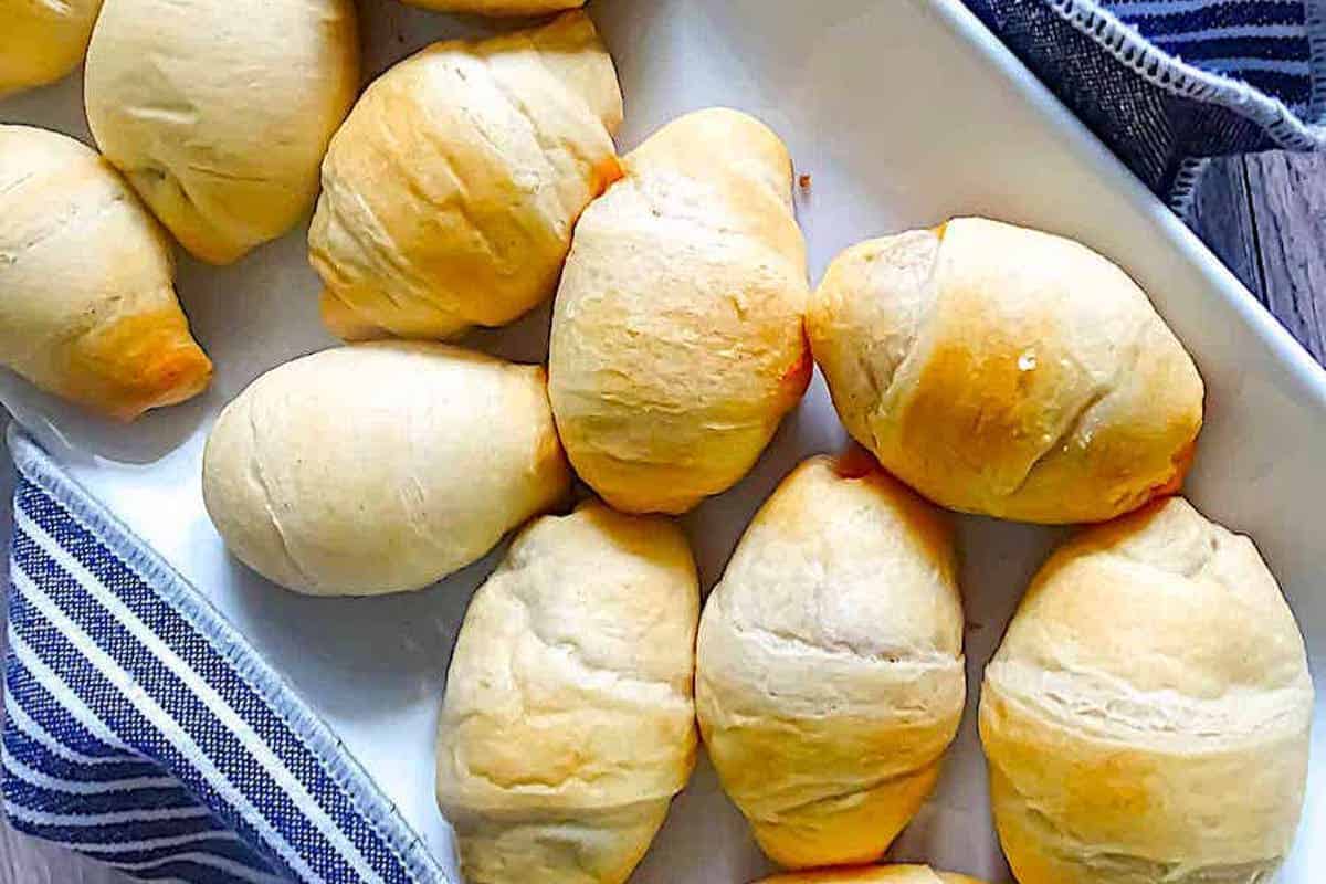A tray of golden-brown mini football pigs in a blanket, arranged neatly on a white baking dish next to a striped cloth napkin, makes the perfect Game Day Party centerpiece. These delicious bites are a must-have for any football-themed recipe collection, sure to be the ultimate touchdown treats!.