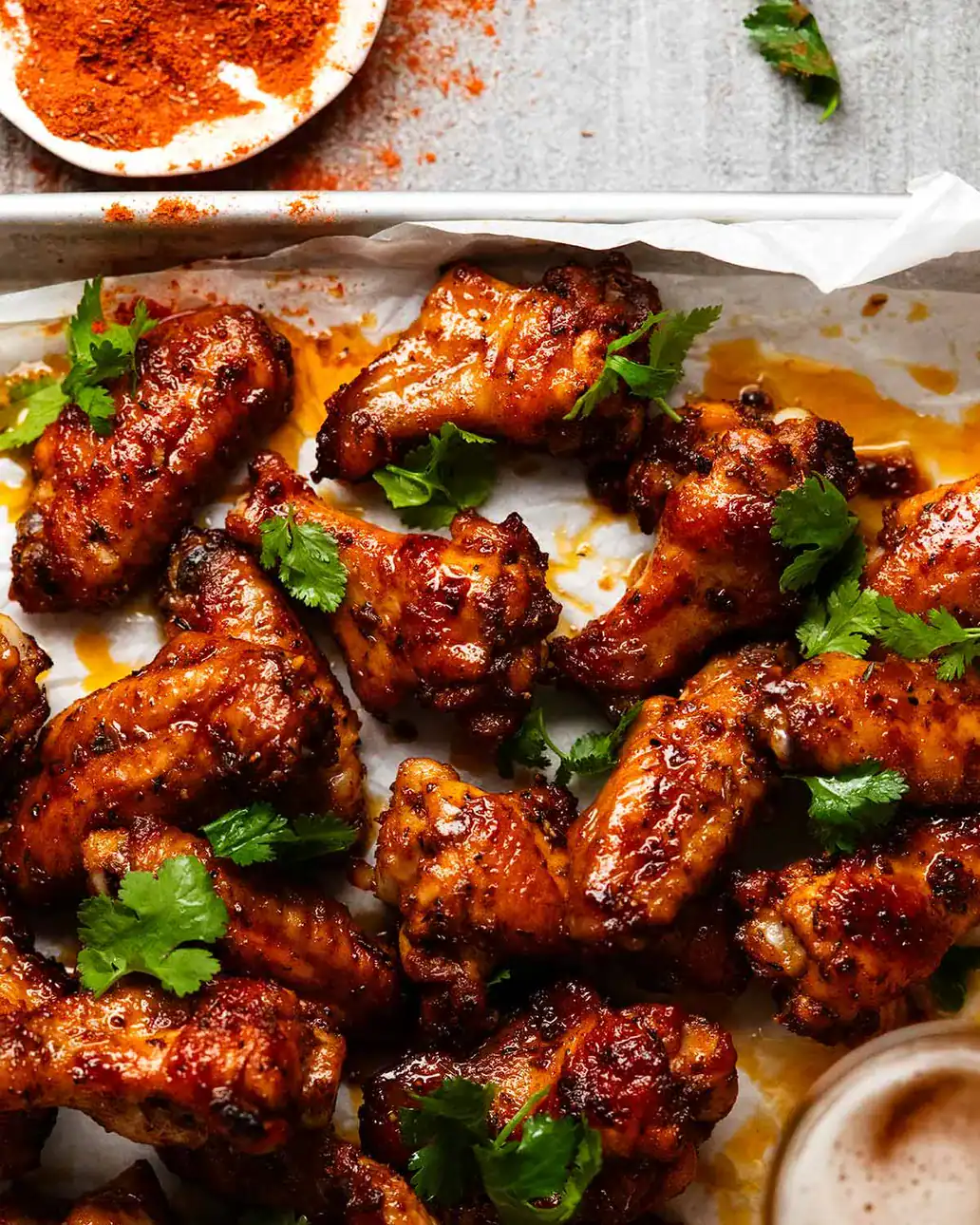 Baked chicken wings coated in a dark glaze, garnished with fresh cilantro, arranged on parchment paper for a finger lickin treat—with a dish of red seasoning and cilantro leaves in the background, perfect for those searching for irresistible wing recipes.
