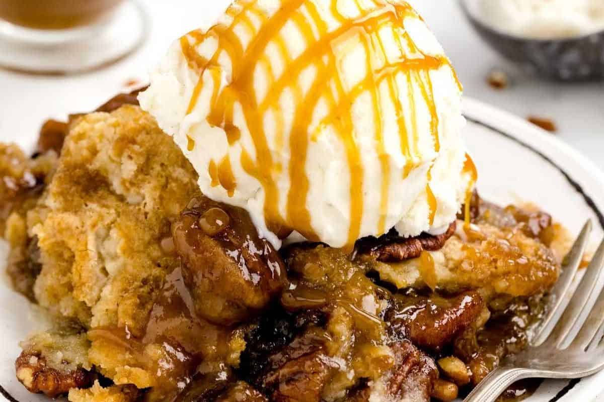 Close-up of a slice of pecan dump cake, served on a white plate and topped with vanilla ice cream and caramel drizzle.