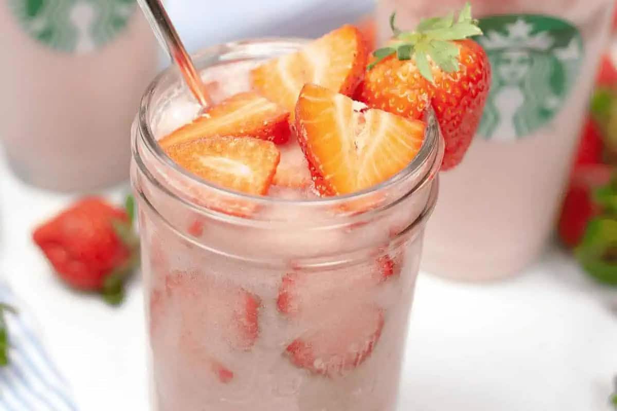 A glass jar filled with iced pink drink, sliced strawberries, and a metal straw sits in front of a Starbucks cup—perfect for those wanting to recreate homemade Starbucks drinks at home.
