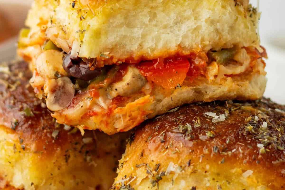 A close-up of two slider buns, the top one cut in half to show a pizza-like filling with sauce, cheese, olives, and mushrooms. Both buns are topped with dried herbs.