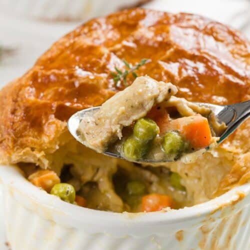 A spoon holds a serving of chicken pot pie with peas, carrots, and chicken above a ramekin topped with golden puff pastry—an ultimate comfort food classic.