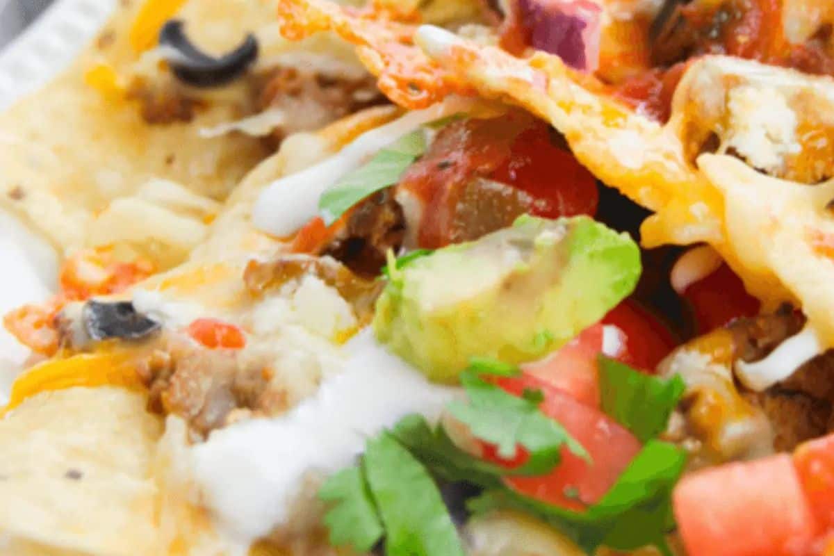 Close-up of loaded nachos topped with melted cheese, avocado, tomatoes, black olives, ground beef, cilantro, and drizzled with white sauce—perfect for a Game Day Party.