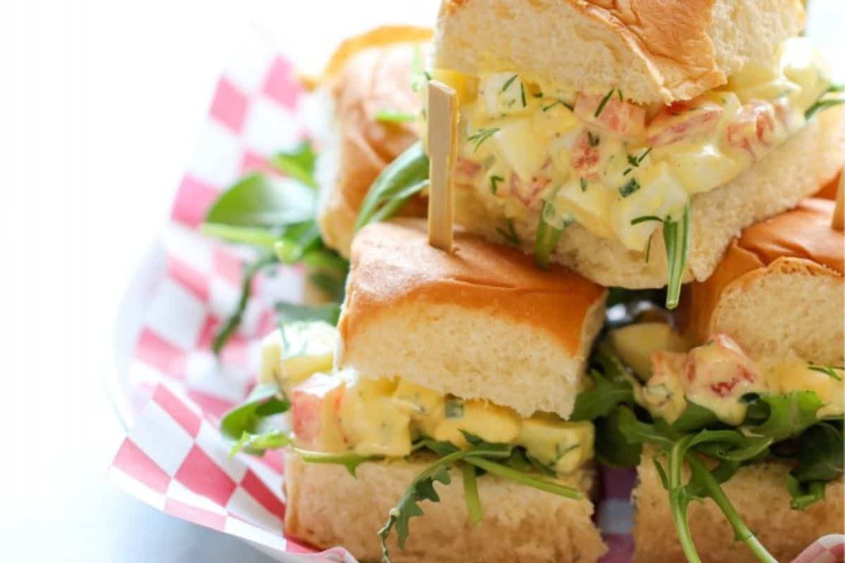 A stack of small sandwiches filled with creamy egg salad and green leafy vegetables, with one sandwich held in place by a wooden pick. The sandwiches are on a plate lined with red and white checkered paper.