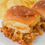 Close-up of a sloppy joe slider with cheese on a sesame seed bun, served on a white plate with a side of potato chips.