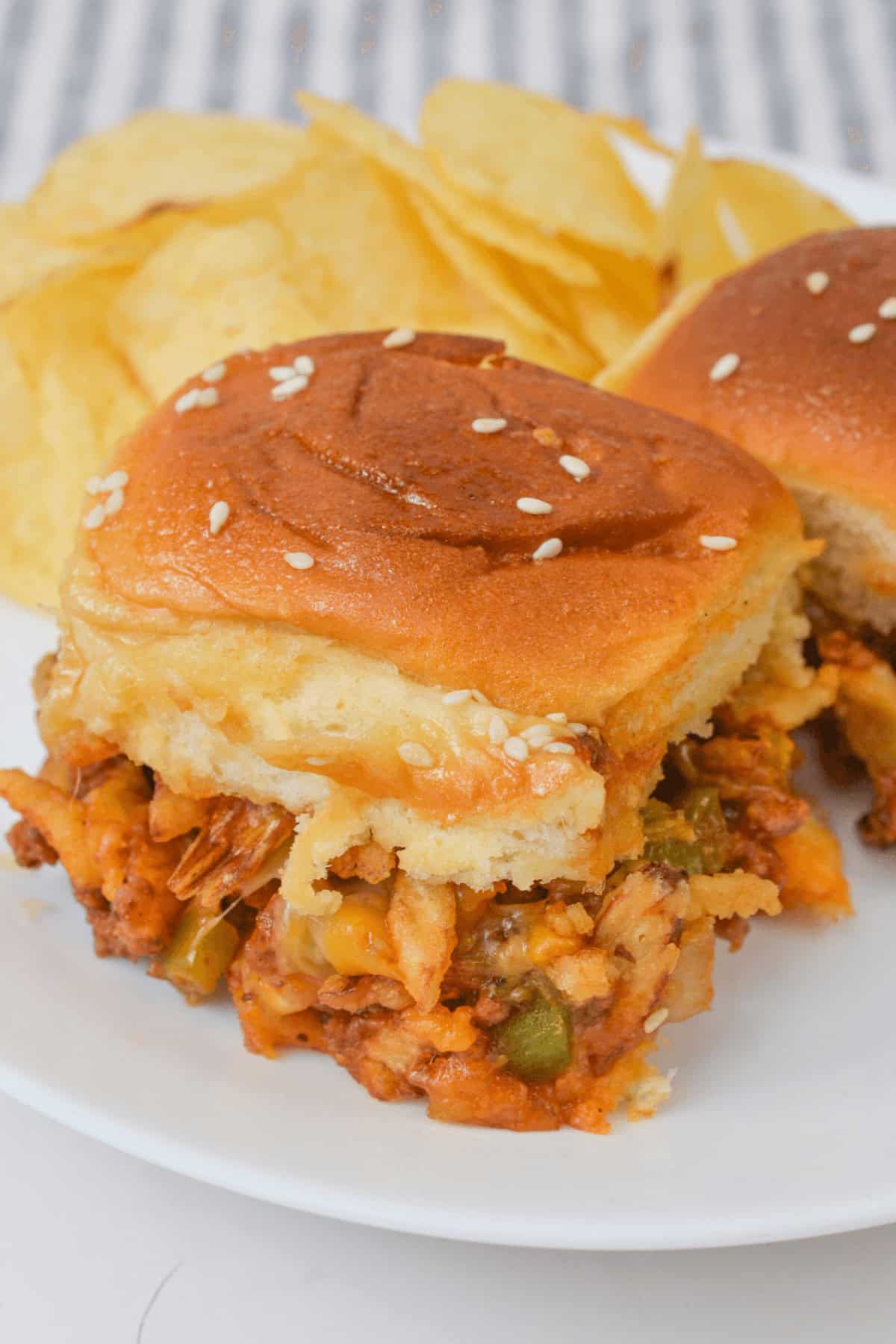 Close-up of a sloppy joe slider with cheese on a sesame seed bun, served on a white plate with a side of potato chips.