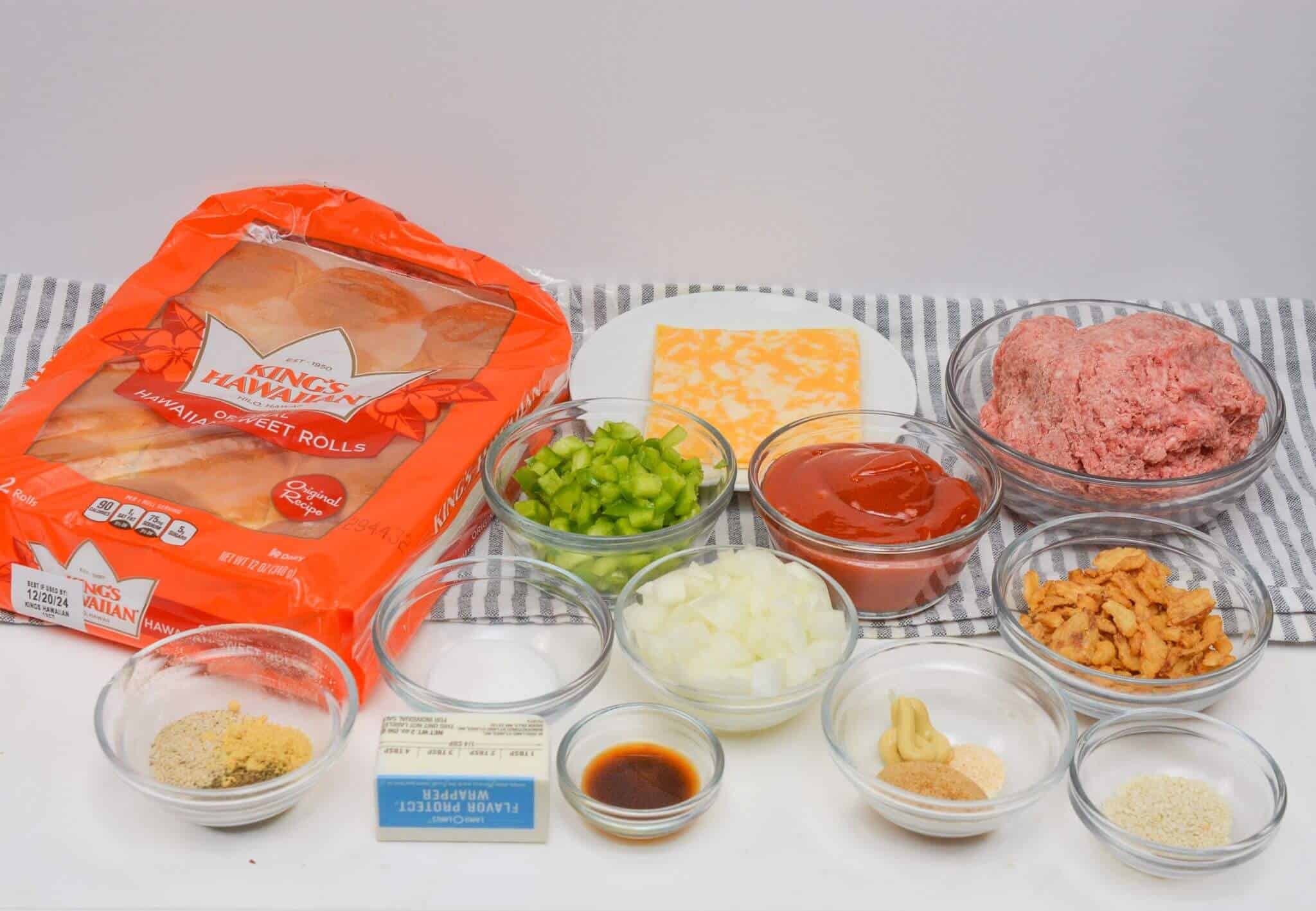 Ingredients for a Sloppy Joe Slider recipe are elegantly arranged on a striped cloth: hamburger rolls, cheese slices, ground beef, diced green peppers and onions, ketchup, and various seasonings in bowls.
