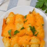 A plate of meal topped with melted cheese and garnished with parsley, placed on a white plate. A fork and parsley leaves are in the background.