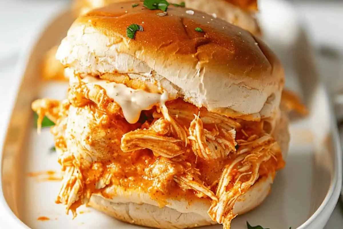 A shredded buffalo chicken slider with sauce on a bun, garnished with small green herbs, served on a white plate—perfect for Game Day.