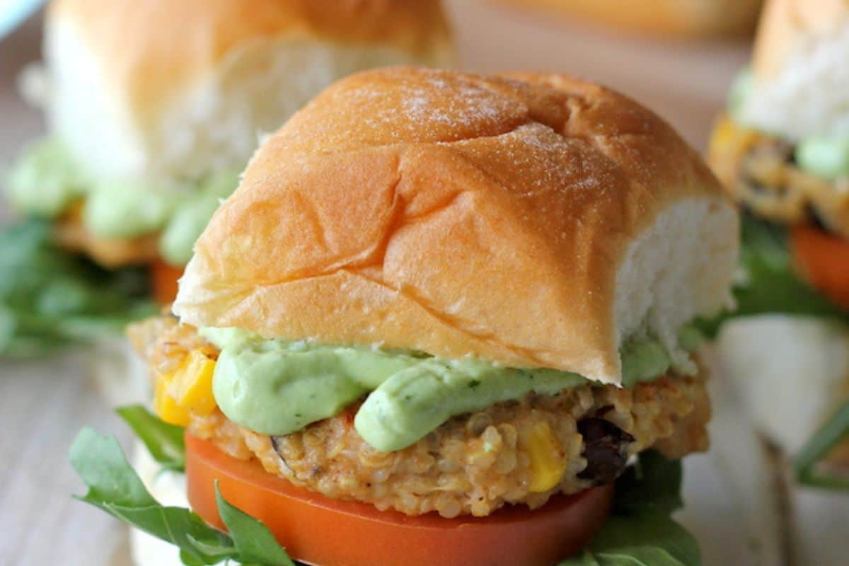 A close-up of a slider with a quinoa patty, a slice of tomato, and green leafy vegetables, topped with a dollop of light green avocado cream sauce. Another slider is visible in the background.