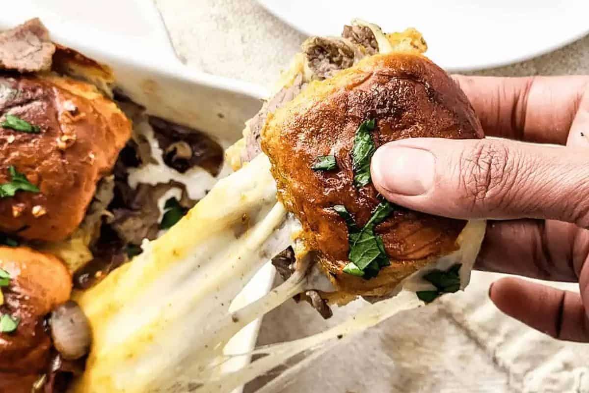 A hand holds a slider bun filled with steak and melted white cheese, which stretches in long strings from the sandwich. Sprinkled green herbs are visible on top of the bun