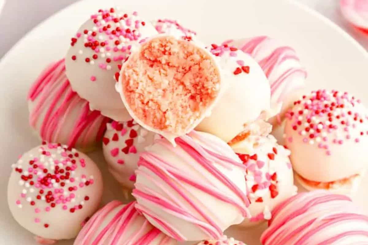 Plate of pink and white cake balls, adorned with drizzles and sprinkles, offering a peek inside one ball. These delightful bites make perfect Little Sweethearts or Valentine Treat Ideas to charm your loved ones.