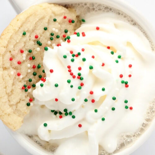 A cup of hot chocolate topped with whipped cream and red and green sprinkles, garnished with a sugar cookie on the rim—just like Starbucks drinks you can recreate at home.