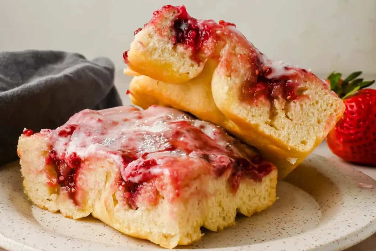 Two pieces of sweet bread topped with a berry glaze are stacked on a plate, perfect for lazy brunch or relaxed weekend mornings. A strawberry and a gray napkin appear in the background.