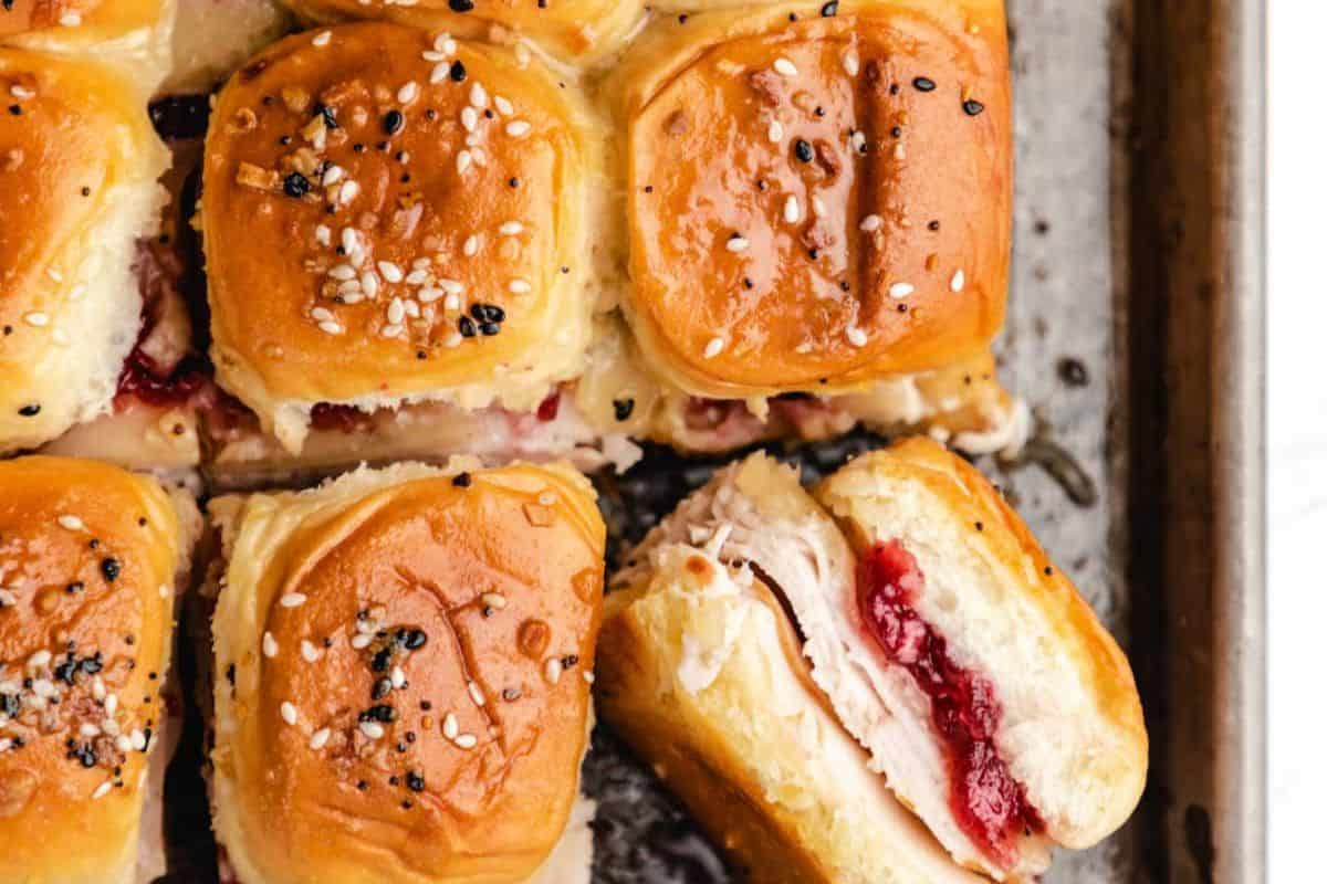An overhead shot of several slider buns, some topped with black and white sesame seeds. One slider is pulled apart, revealing slices of turkey and a layer of red cranberry sauce inside.