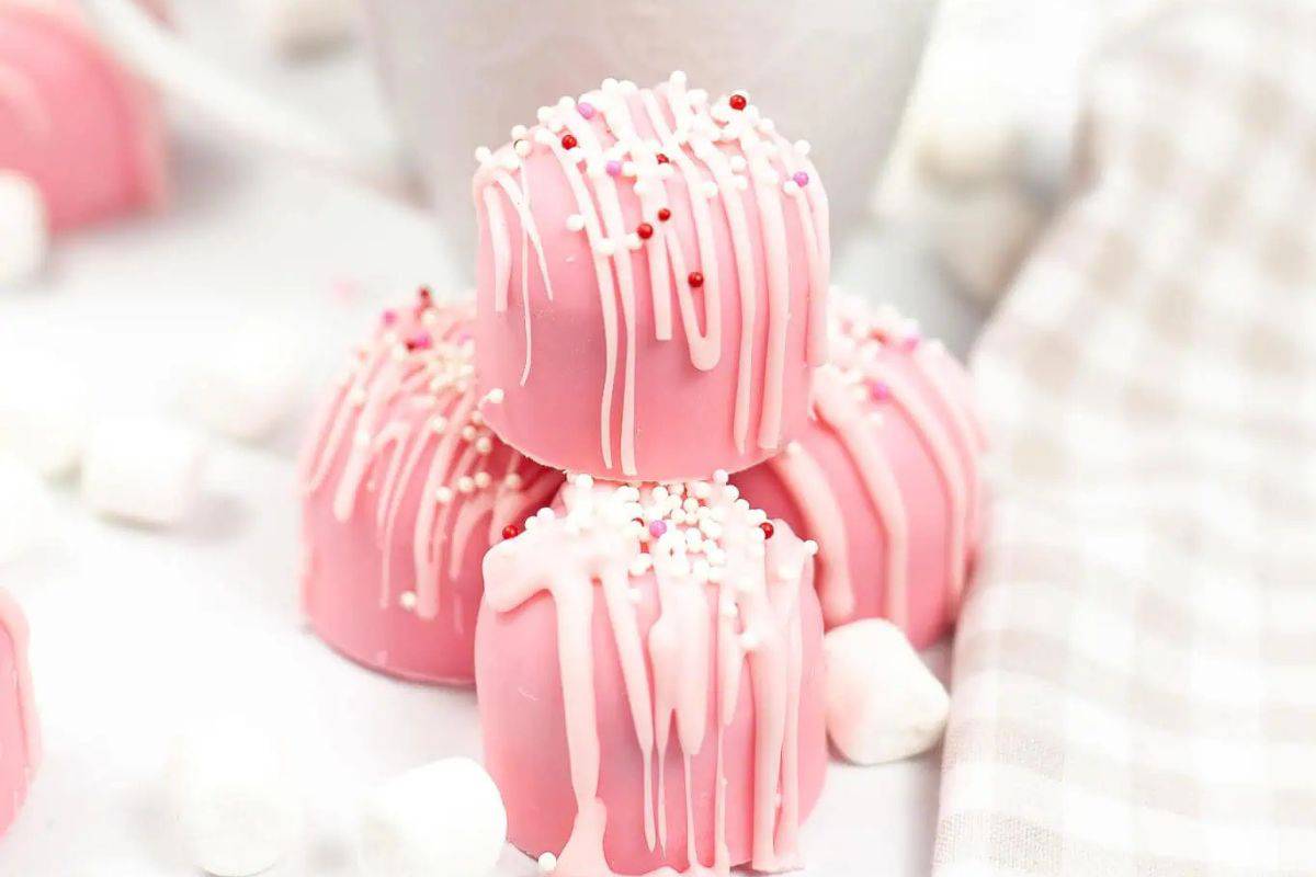Three pink chocolate-covered treats with white icing and sprinkles, whimsically stacked on a table near a checked cloth and mini marshmallows. These delightful confections are perfect creative Valentine treat ideas for your little sweethearts.