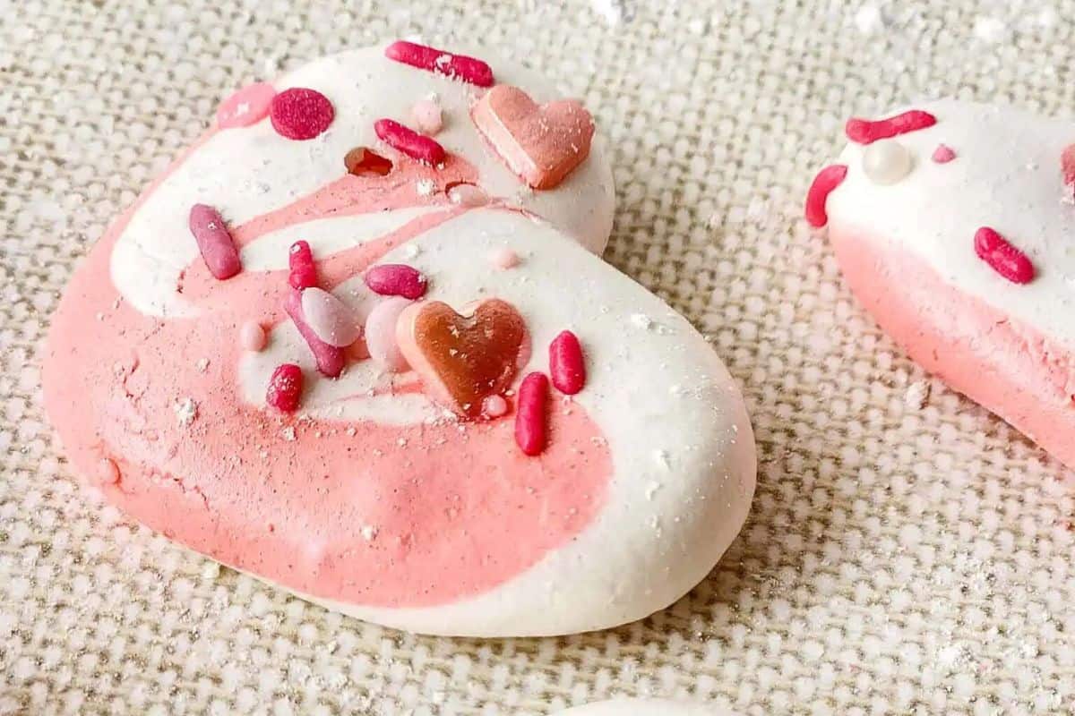 A heart-shaped cookie with pink and white icing, creatively topped with various sprinkles, including heart shapes, on a textured surface. It's a delightful Valentine treat idea for your little sweethearts.