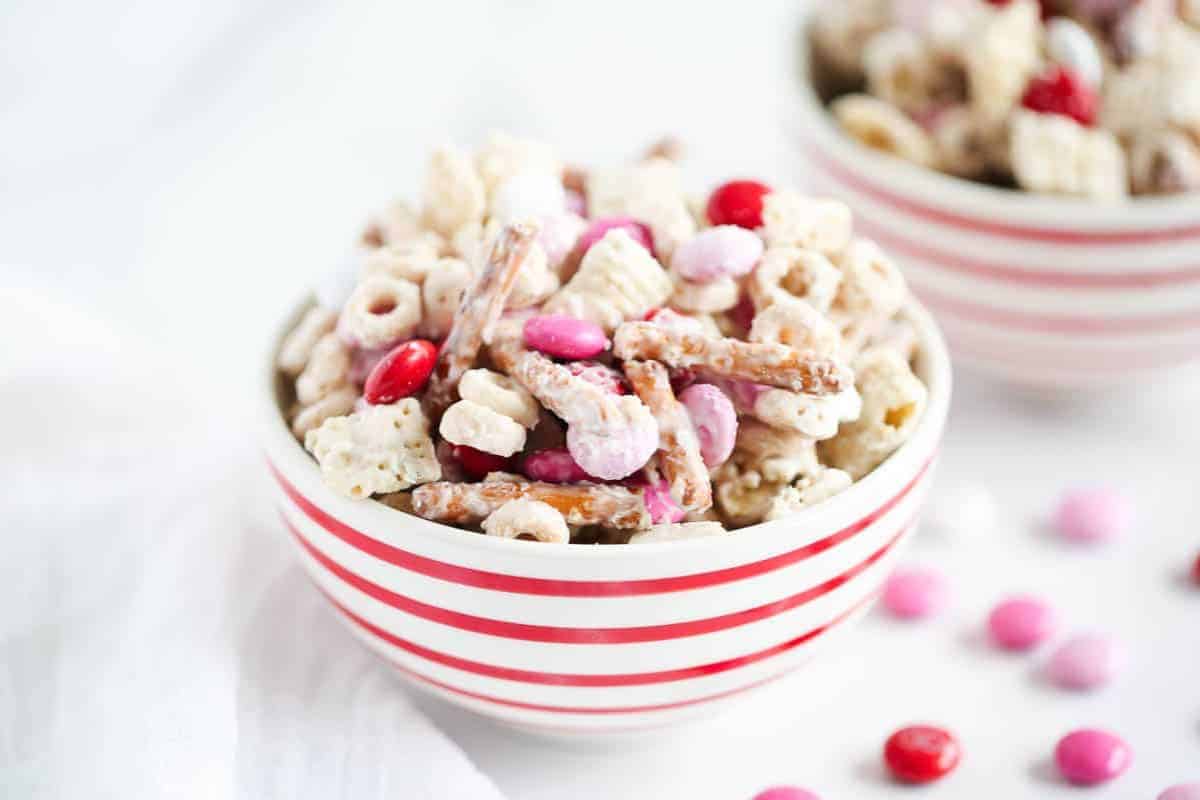 A bowl of snack mix with cereal, pretzel sticks, and pink and red candies sits in a red and white striped bowl—perfect for Creative Valentine treat ideas.