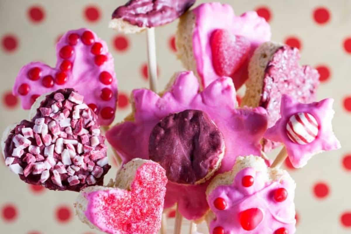 An assortment of heart-shaped cookies and treats on sticks, covered in pink and red icing with various decorations, offers delightful inspiration for Valentine Treat Ideas. These creative Valentine treats are displayed against a playful polka dot background.