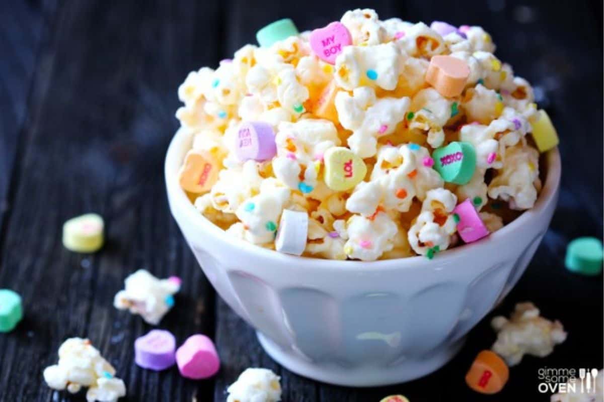 A white bowl of popcorn mixed with colorful, heart-shaped candies sits on a dark wooden surface, offering a creative twist on Little Sweethearts—a delightful treat perfect for Valentine celebration ideas.