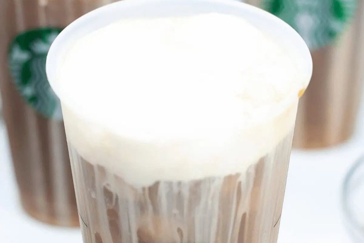 A close-up of a plastic cup filled with iced coffee topped with cold foam, with other Starbucks drinks blurred in the background—perfect inspiration to recreate at home for your next DIY Starbucks treat.