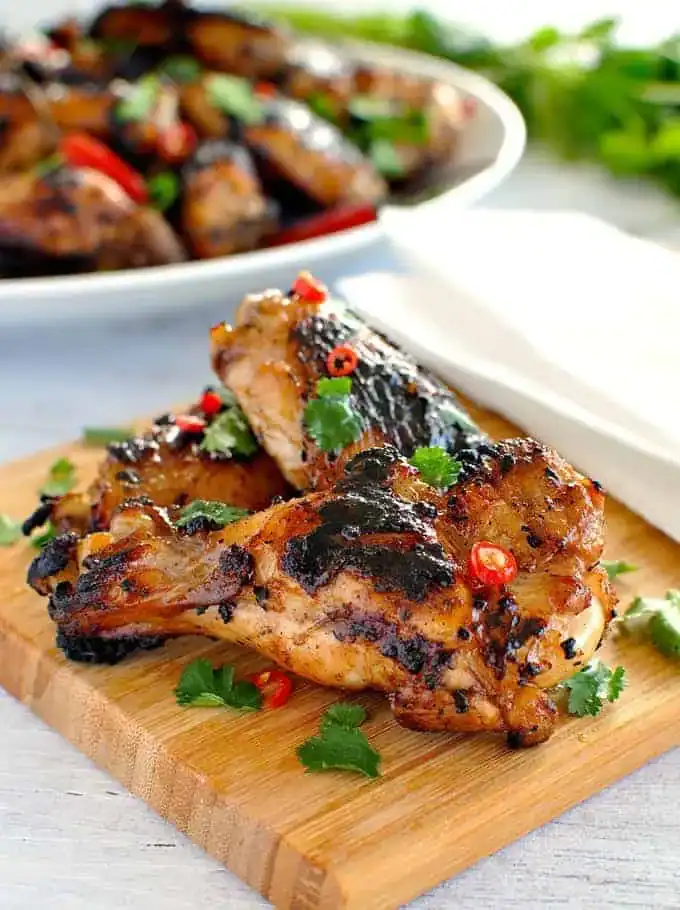 Two grilled chicken pieces with char marks, reminiscent of classic finger lickin’ wing recipes, are garnished with cilantro and sliced red chili on a wooden cutting board. More chicken wings are visible blurred in the background.