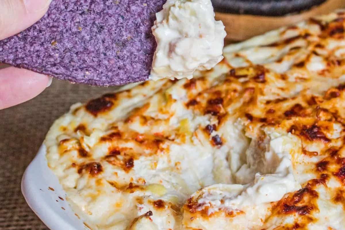 A hand dips a blue corn tortilla chip into a dish of baked, cheesy, golden-brown dip—perfect for those who love copycat recipes to make at home.