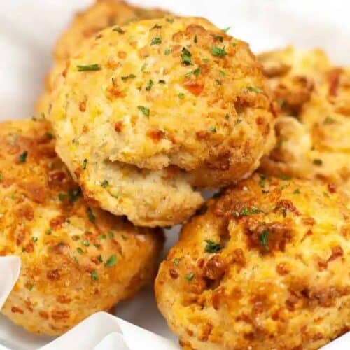 Several golden-brown cheddar biscuits with parsley garnish are stacked on white parchment paper in a bowl, perfect for those searching for Red Lobster copycat recipes to make at home.