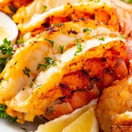 Perfect for special dinners or cozy nights, these grilled lobster tails are garnished with chopped parsley and served with lemon wedges and melted butter on a white plate. A standout among lobster recipes!.