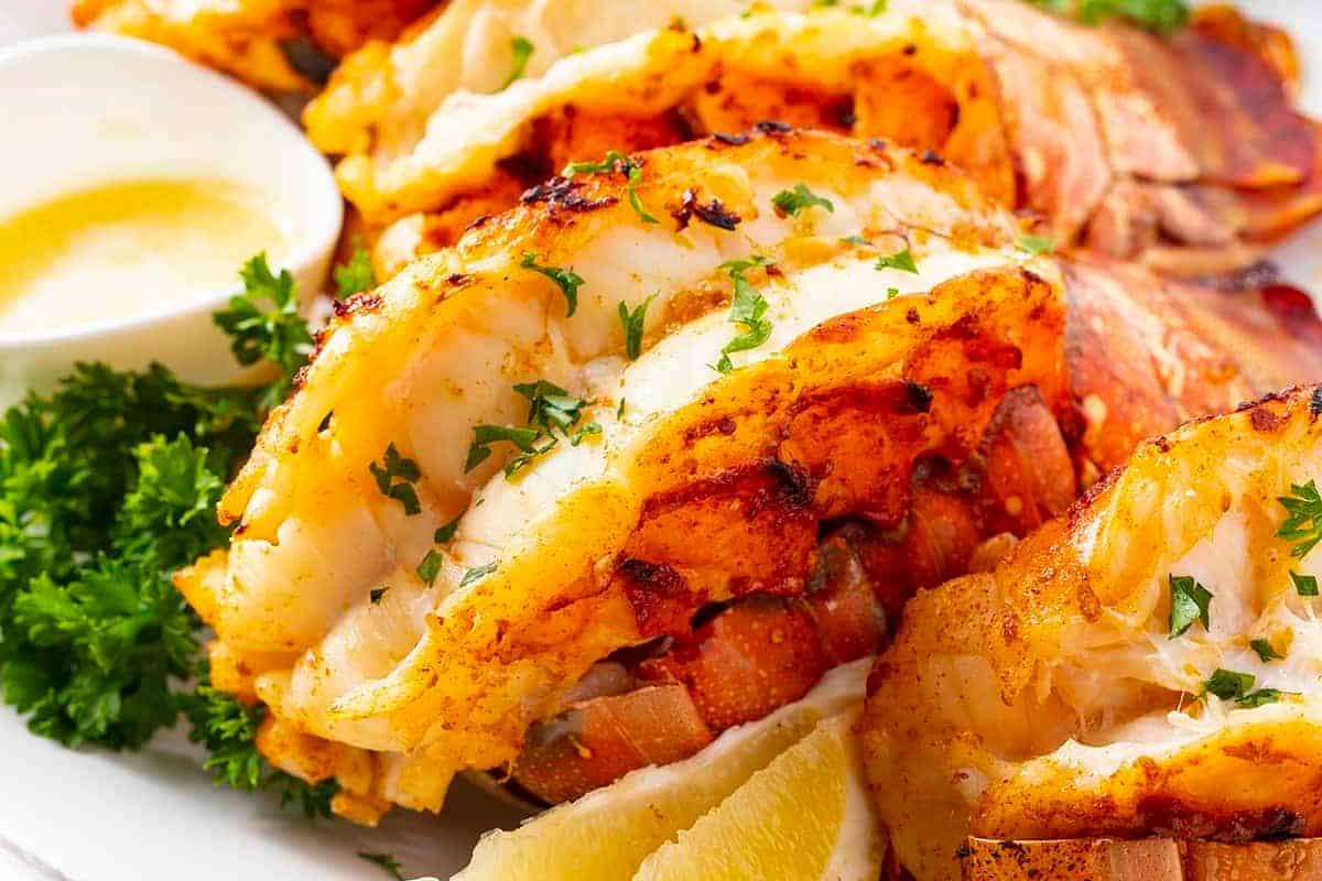 Perfect for special dinners or cozy nights, these grilled lobster tails are garnished with chopped parsley and served with lemon wedges and melted butter on a white plate. A standout among lobster recipes!.