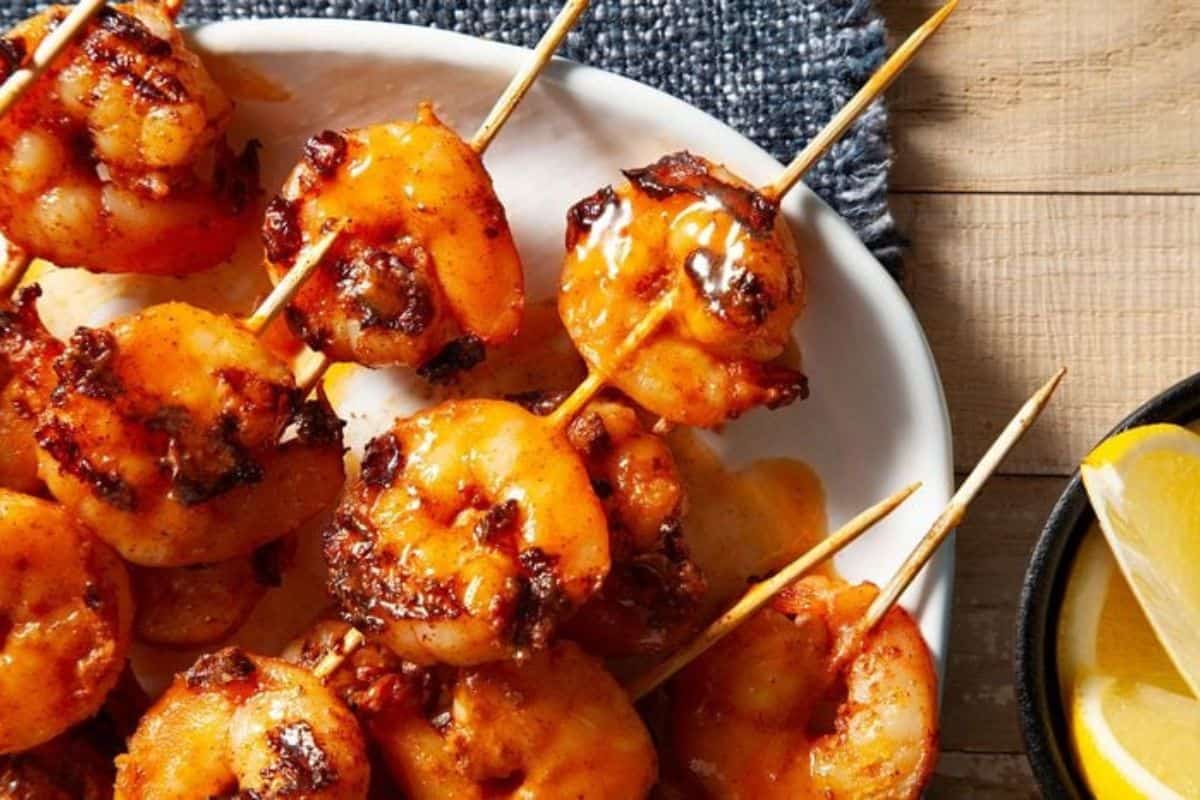 Grilled shrimp skewers on a white plate with a blue napkin, next to lemon wedges in a black bowl on a wooden surface—an easy Red Lobster copycat recipe you can make at home for a delicious seafood feast.