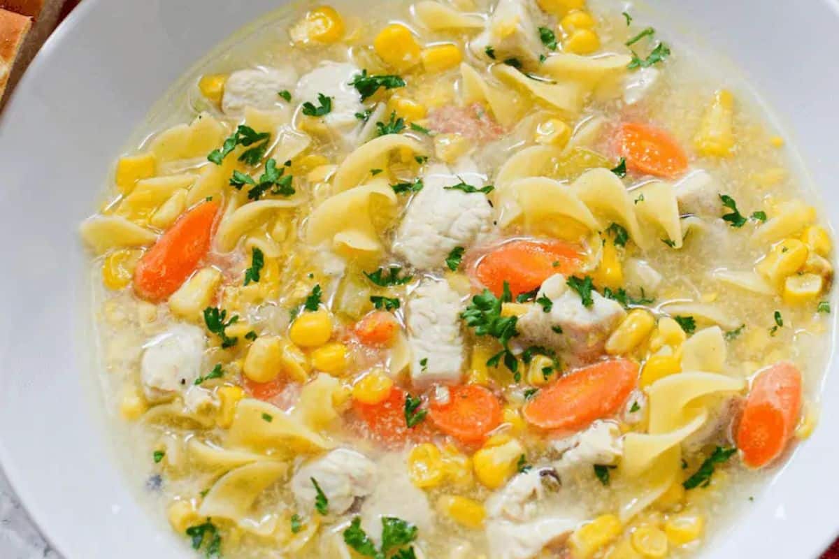A bowl of chicken noodle soup with egg noodles, corn, carrots, chopped parsley, and chunks of chicken in broth—a classic among comforting soups.