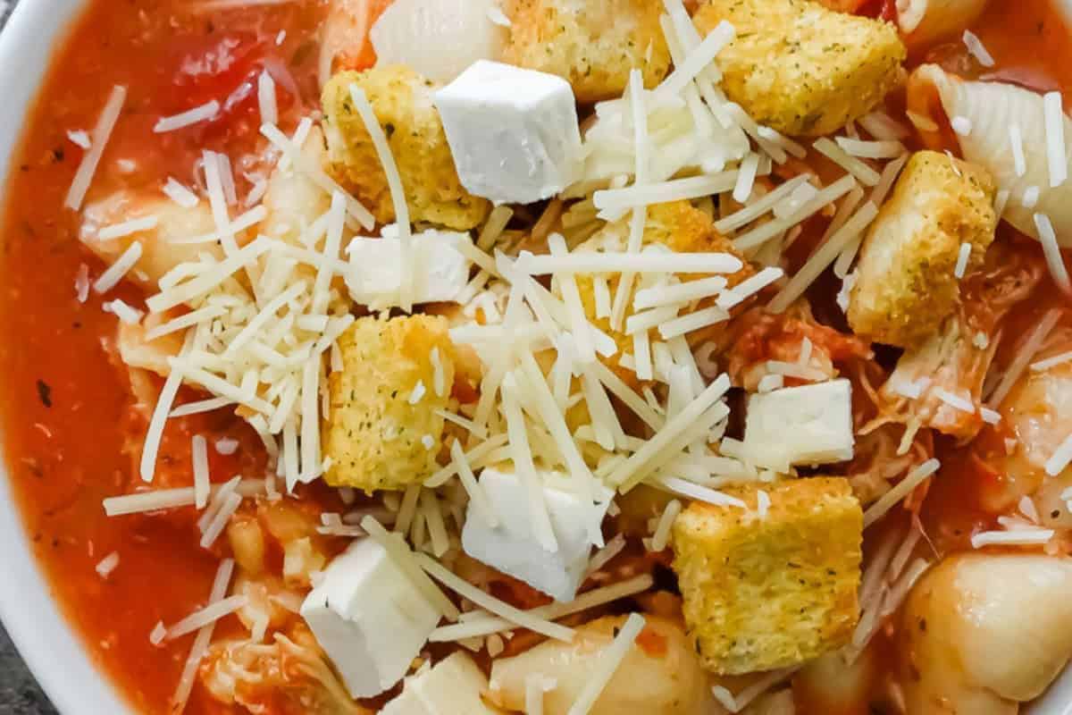A bowl of tomato-based pasta soup topped with shredded cheese, croutons, and small cubes of white cheese—perfect for those who love family friendly soups.