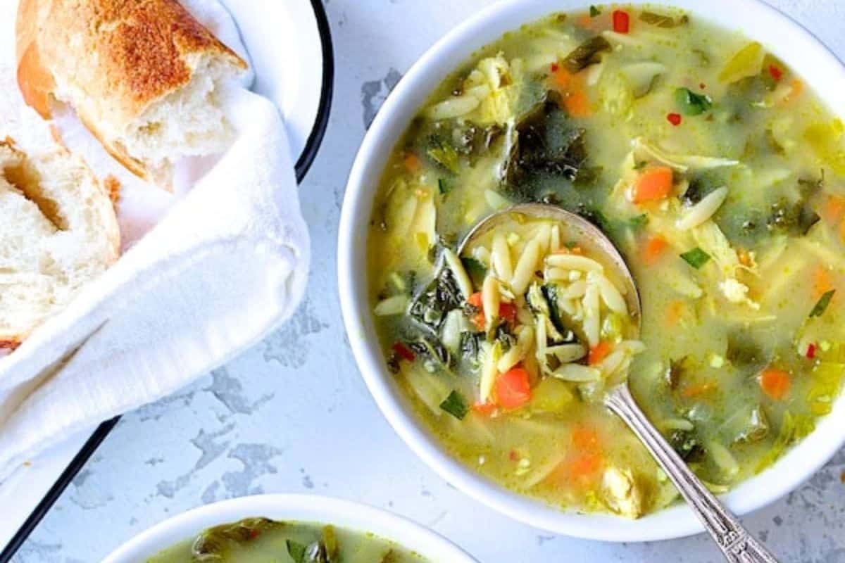 A bowl of comforting soup with orzo pasta and greens, served with a spoon, sits beside a plate of sliced bread wrapped in a white cloth—perfect to warm your soul on a chilly day.