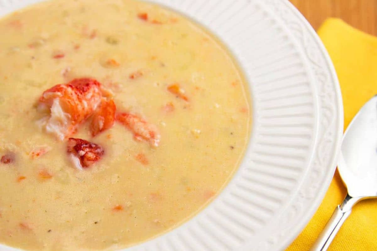 A bowl of creamy lobster bisque with visible lobster pieces, served in a white dish next to a spoon and a yellow napkin—the perfect addition to cozy nights or special dinners.