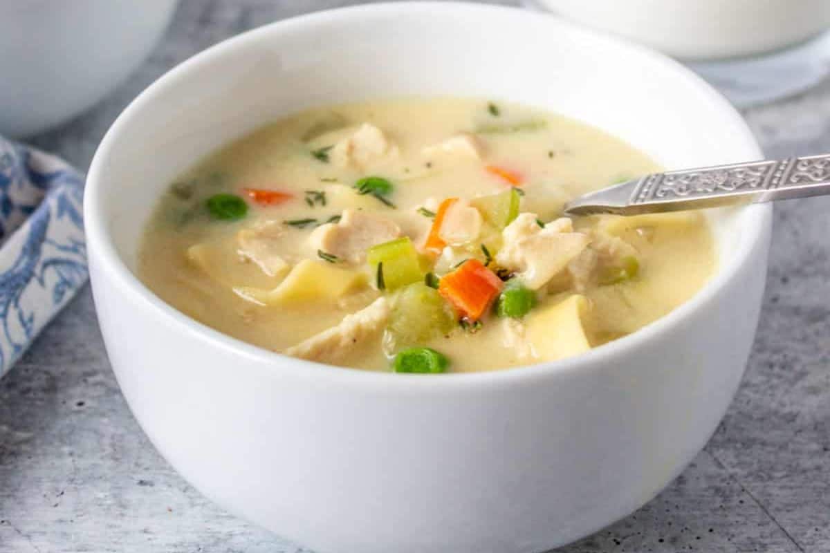 A white bowl filled with creamy chicken soup containing chunks of chicken, peas, carrots, and potatoes, with a spoon resting inside—one of those comforting soups sure to warm your soul.