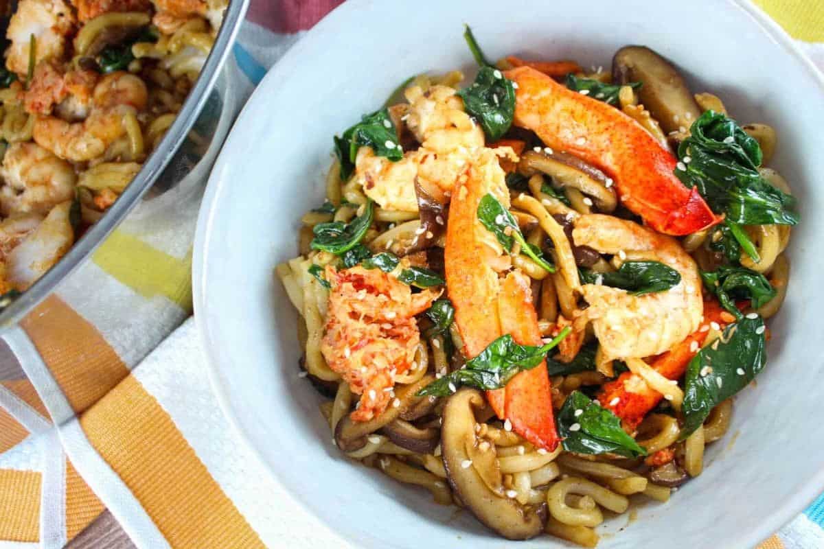 A bowl of stir-fried udon noodles with shrimp, lobster, spinach, mushrooms, and sesame seeds—perfect for cozy nights or special dinners.