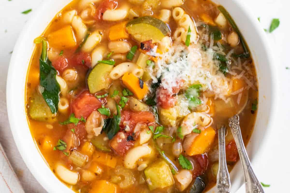 A bowl of vegetable soup with macaroni, zucchini, tomatoes, carrots, beans, greens, and a sprinkle of grated cheese—with two spoons ready to share. This is one of the best family friendly soups with pasta everyone will enjoy.