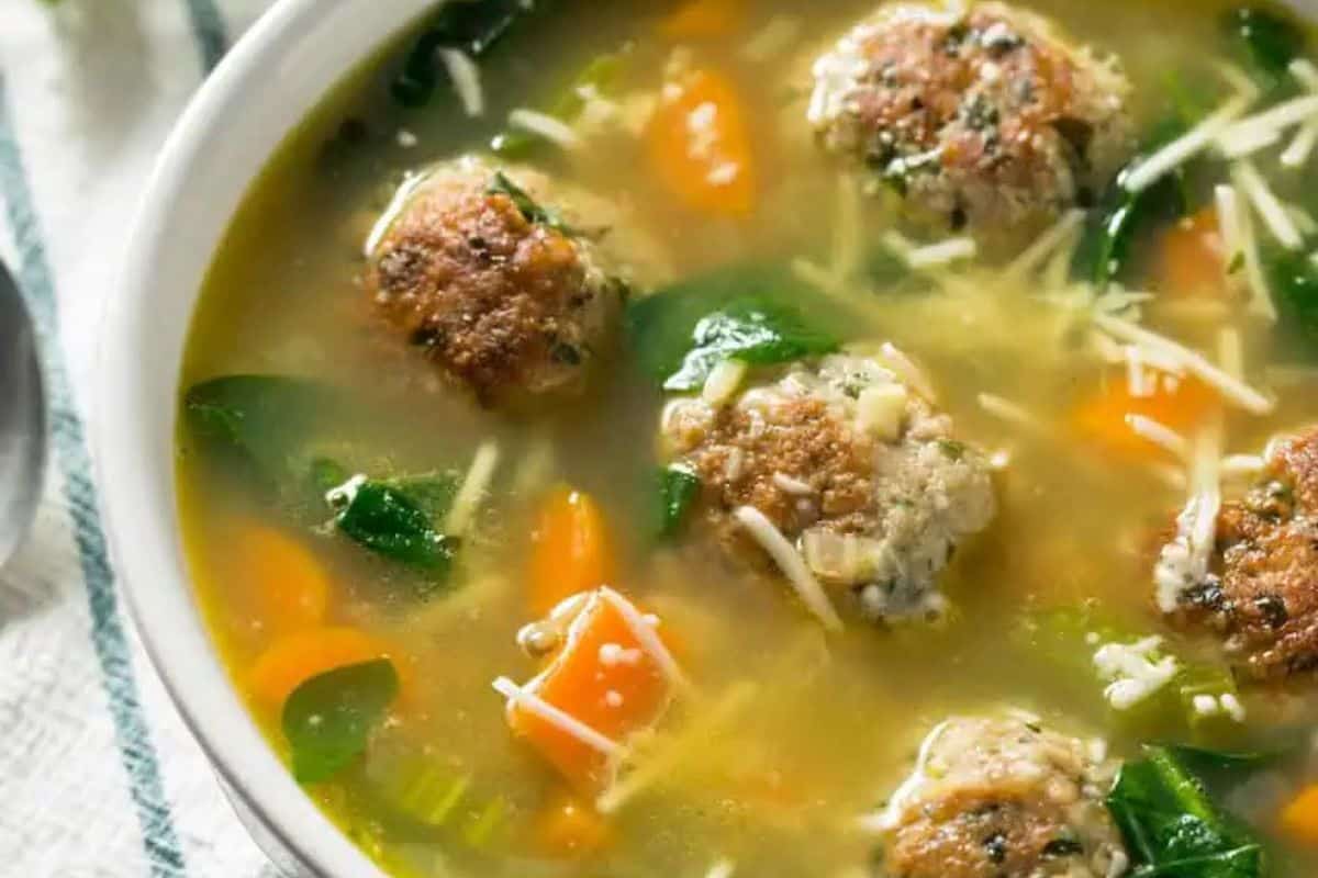 A bowl of family friendly soup with meatballs, chopped carrots, spinach, pasta cubes, and shredded cheese in a clear broth.