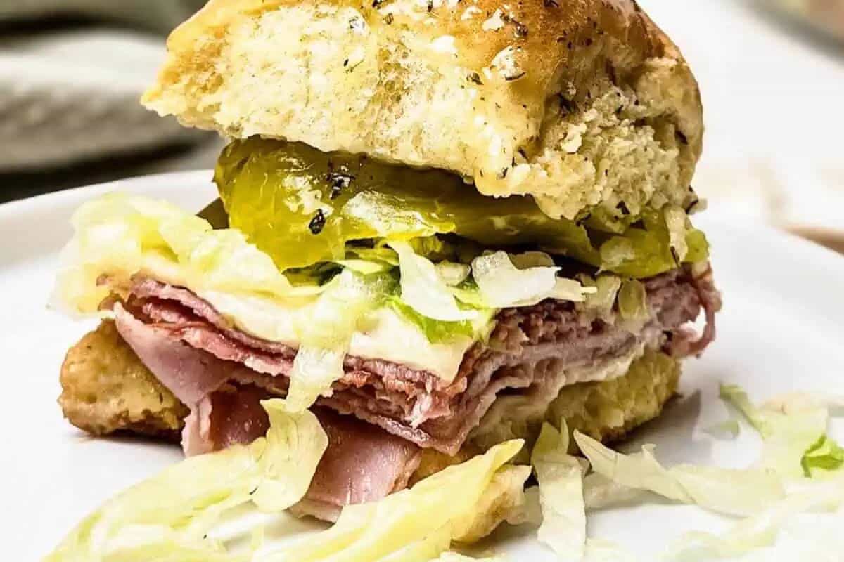 A close-up of an Italian sub slider, showing layers of deli meats, white cheese, shredded lettuce, and pickled peppers spilling out from a seasoned bun.