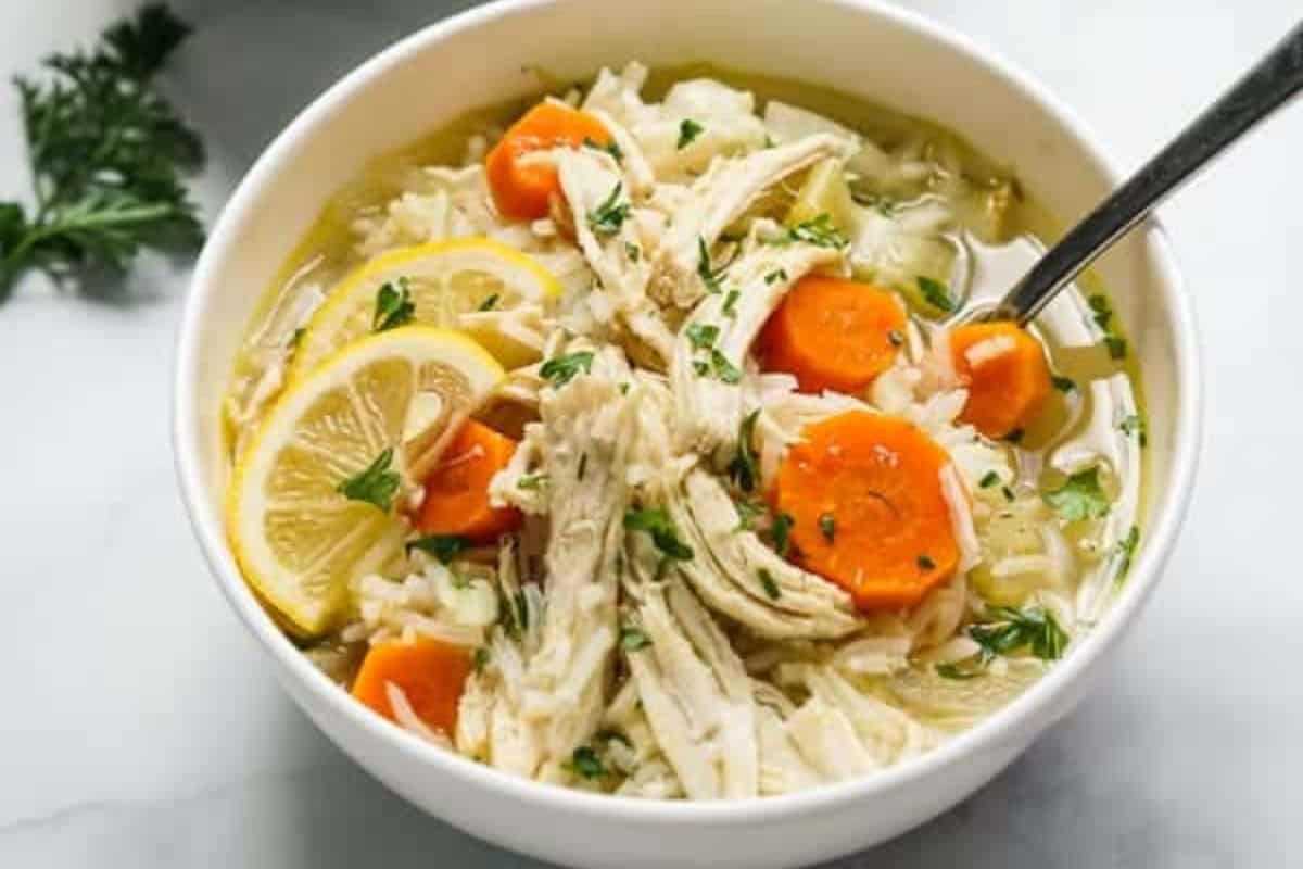 A bowl of chicken soup with shredded chicken, carrot slices, lemon wedges, rice, and herbs, with a spoon resting inside—one of those comforting soups sure to warm your soul.