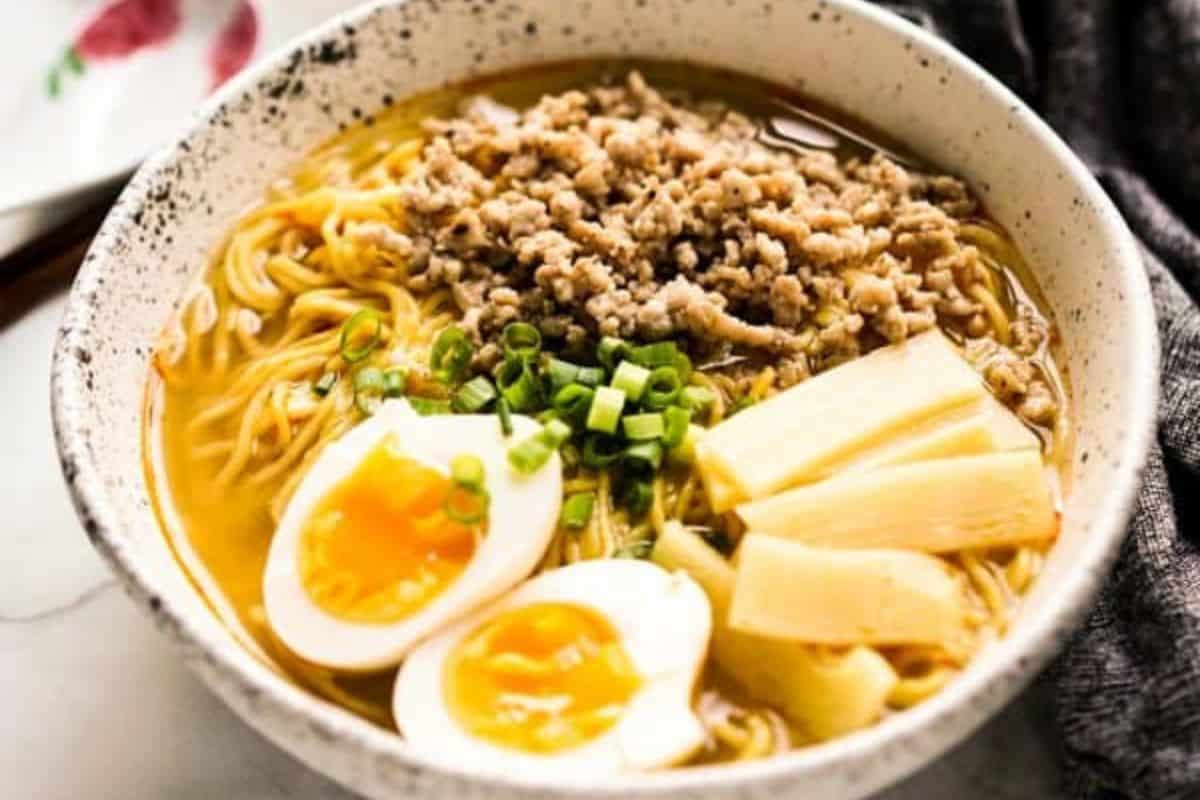 A bowl of ramen noodles topped with ground meat, sliced green onions, bamboo shoots, and two halved soft-boiled eggs in broth—a comforting twist on soups with pasta that's perfect for family friendly soups.