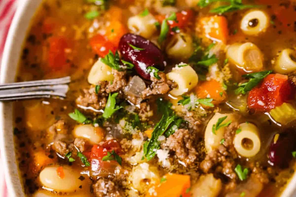 A close-up of a bowl of pasta soup with ground meat, ditalini pasta, kidney beans, diced tomatoes, carrots, celery, and herbs, with a fork resting inside the bowl—a comforting choice among family friendly soups.