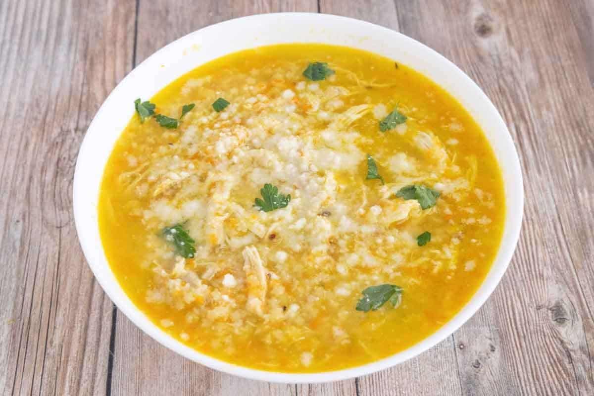 A white bowl of yellow soup with shredded chicken, cheese, and fresh cilantro sits on a wooden surface—one of our favorite family friendly soups that's perfect for any mealtime.