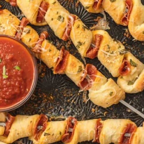 Skewers of twisted dough and pepperoni are arranged on a baking tray with a bowl of marinara sauce for dipping, making them perfect as kid friendly dinner recipes.