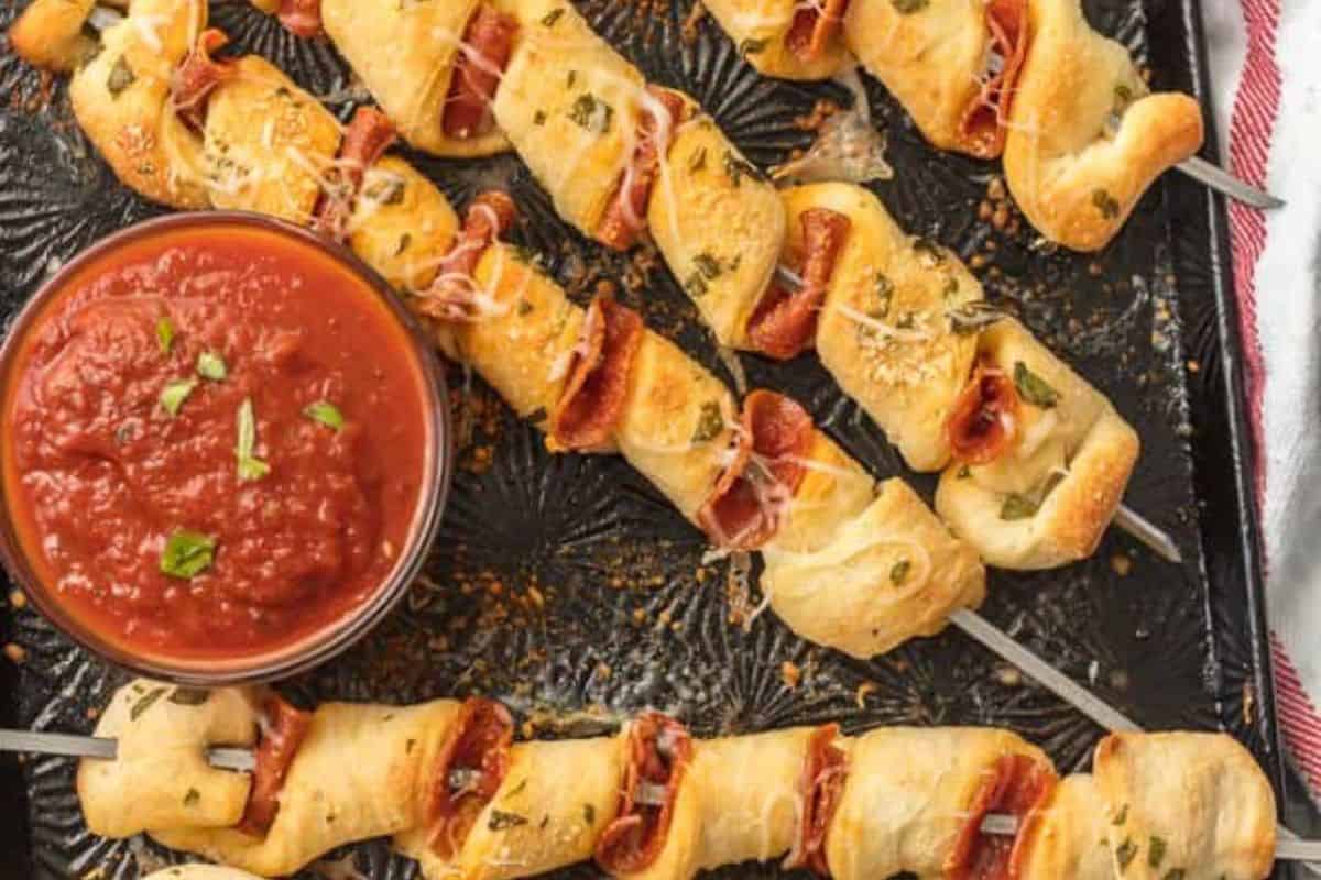 Skewers of twisted dough and pepperoni are arranged on a baking tray with a bowl of marinara sauce for dipping, making them perfect as kid friendly dinner recipes.