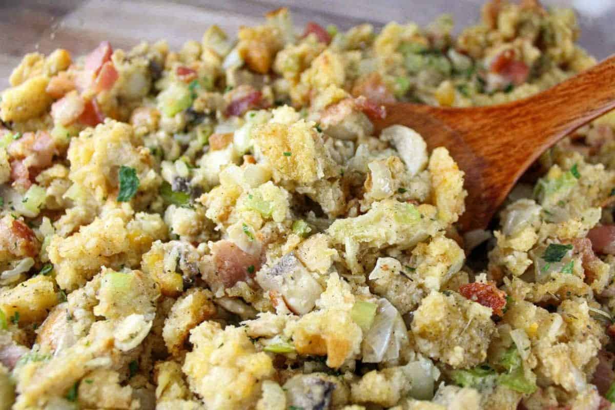 A wooden spoon stirring a dish of cornbread stuffing with visible pieces of bread, vegetables, herbs, and bacon—perfect for those who love copycat recipes to enjoy classic flavors at home.